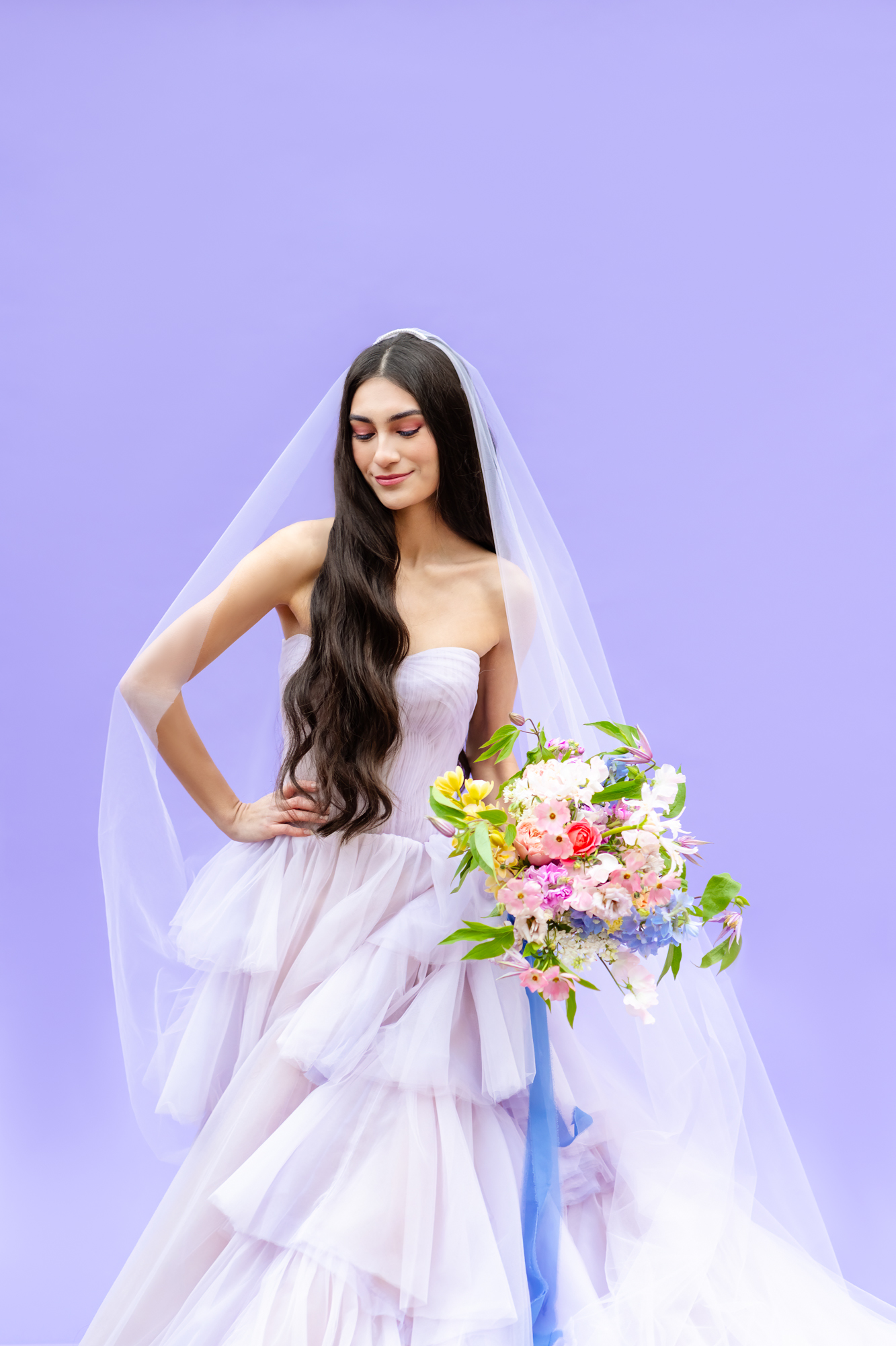A bride wearing a strapless, tiered tulle wedding gown and long veil poses gracefully against a lavender backdrop, holding a vibrant, colorful bouquet of flowers with pink, yellow, blue, and white blooms.A bride wearing a strapless, tiered tulle wedding gown and long veil poses gracefully against a lavender backdrop, holding a vibrant, colorful bouquet of flowers with pink, yellow, blue, and white blooms.