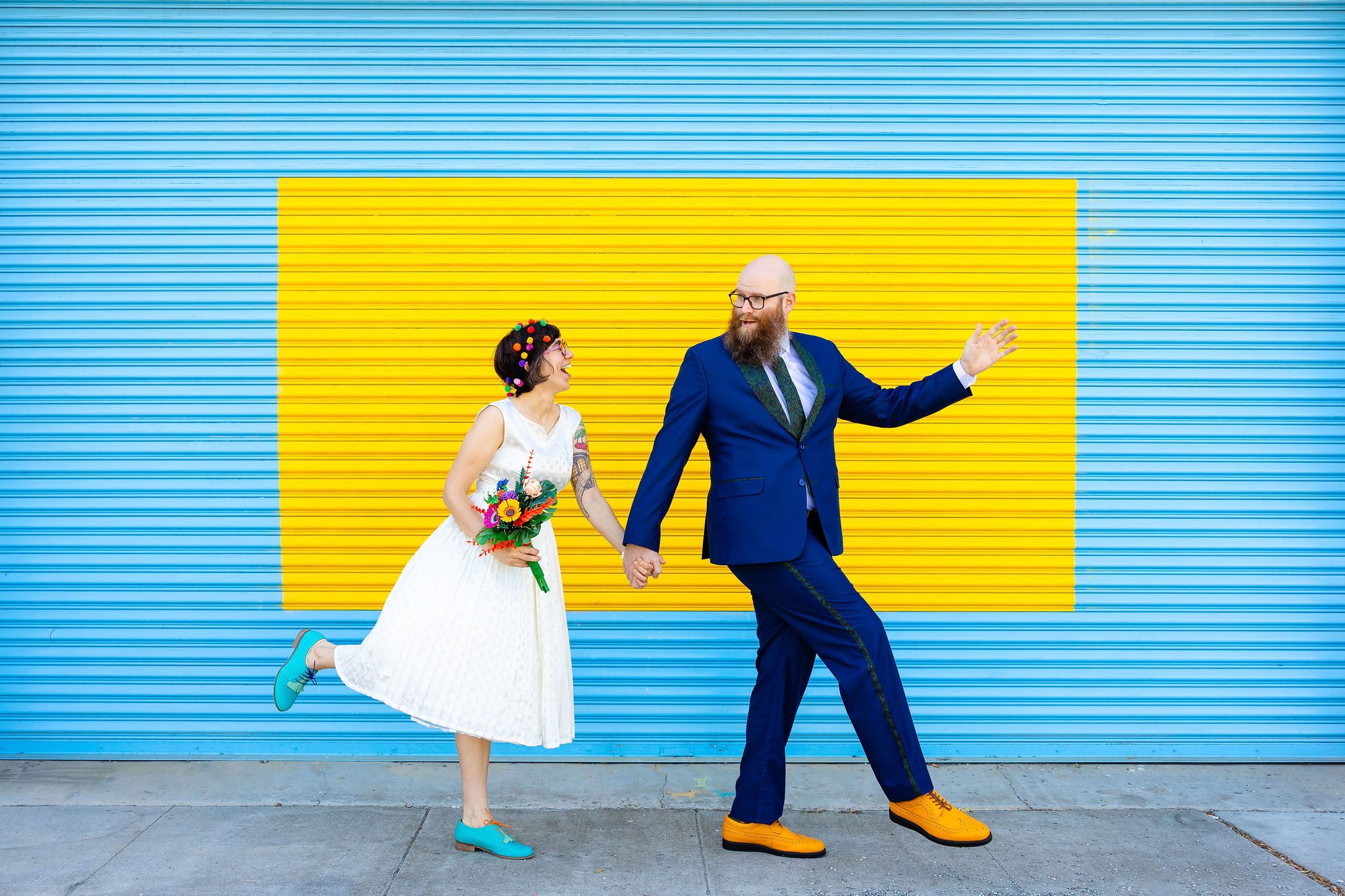 A bride and groom hold hands and walk with a funny pose in front of a blue and yellow wall. The bride is wearing a white vintage wedding dress with blue shoes and holds colorful lego flowers, while the groom wears a blue suit and yellow shoes.