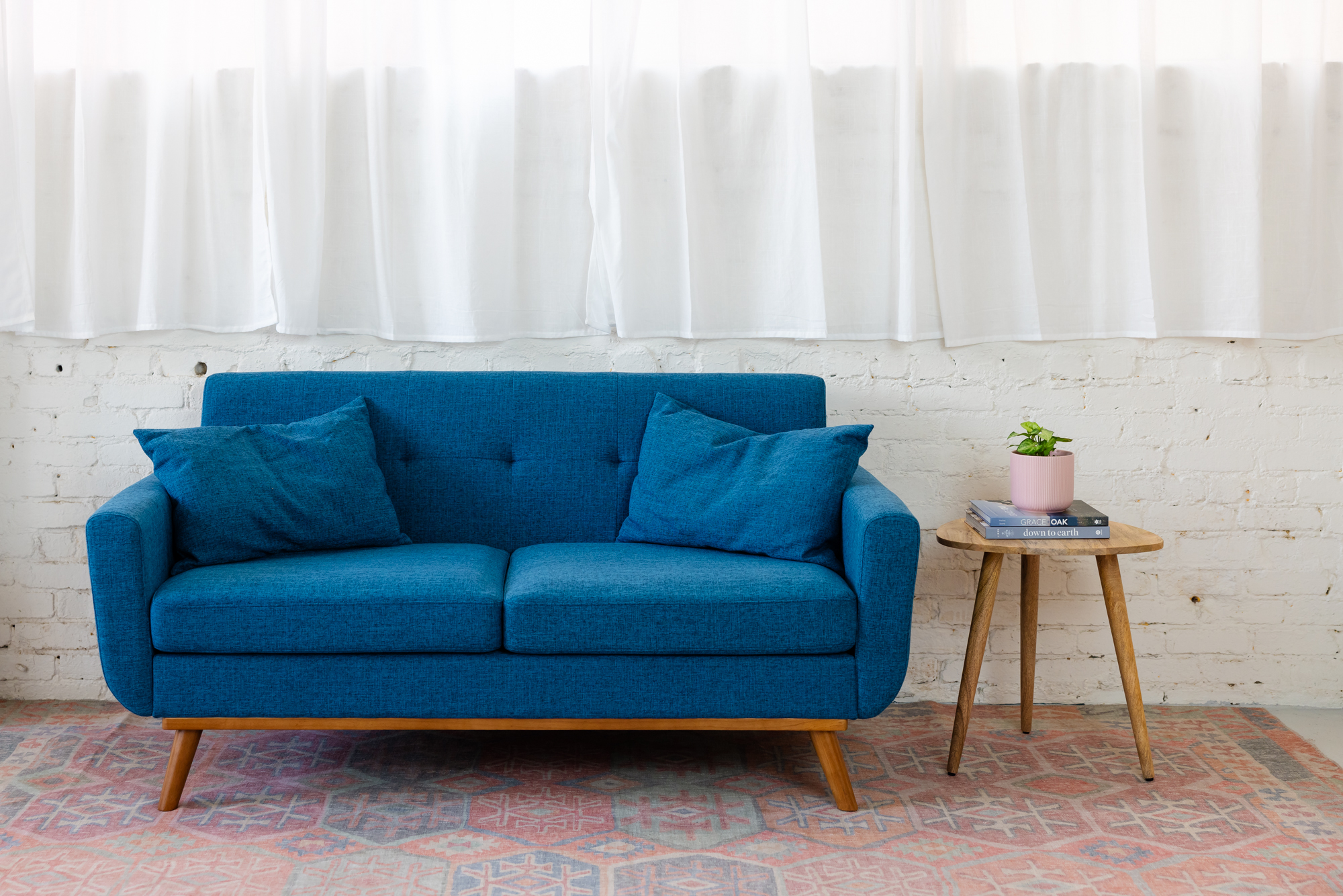 Bright blue sofa in a natural light photography studio with white brick wall, sheer curtains, patterned rug, and small side table with plant in Emeryville studio space