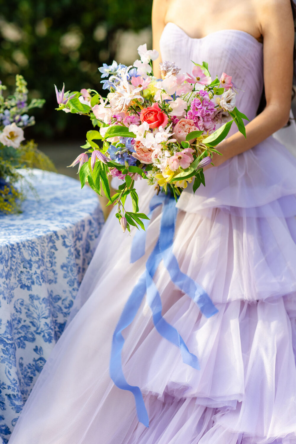 close up of a colorful spring floral bouquet with periwinkle ribbon, held in front of a light lavender tulle wedding gown.