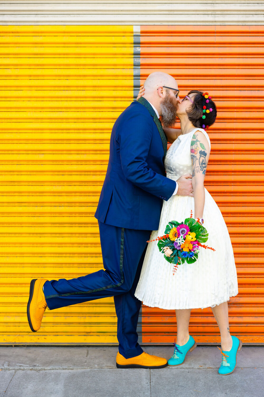 a couple kisses in front of a yellow and orange colored wall. The bride is holding a small colorful bouquet made of legos
