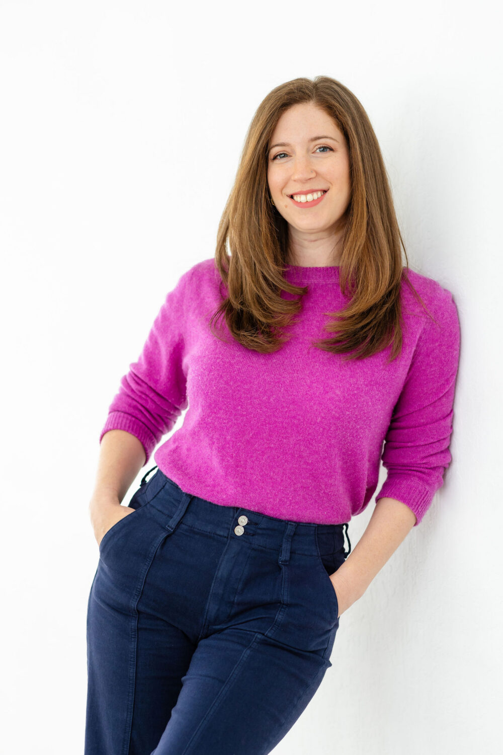 a woman wears a fuchsia sweater and blue pants while leaning on a white wall and looking at the camera