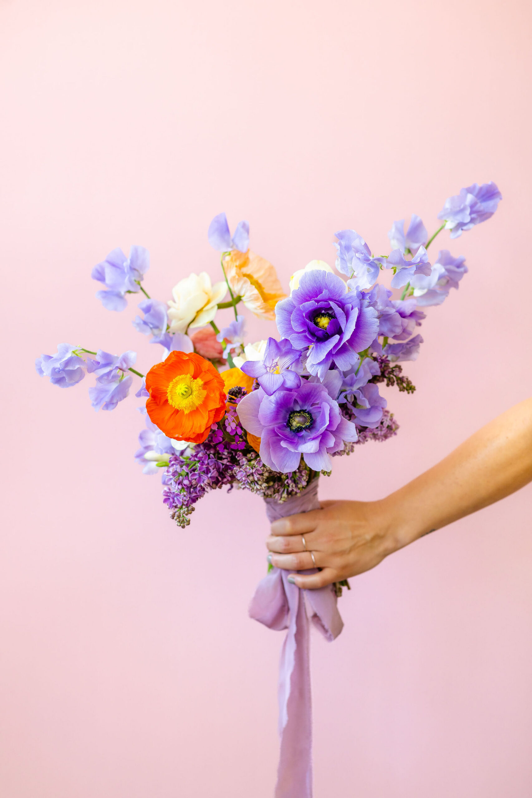 an outstretched hand holds a minimalist wedding bouquet with mostly purple flowers and one orange flower