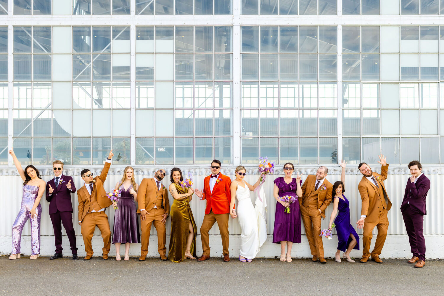 fun photo of a wedding party in front of the bridge yard in Oakland. They're all wearing shades of mustard/khaki or purple. The groom is wearing an orange velvet suit jacket.