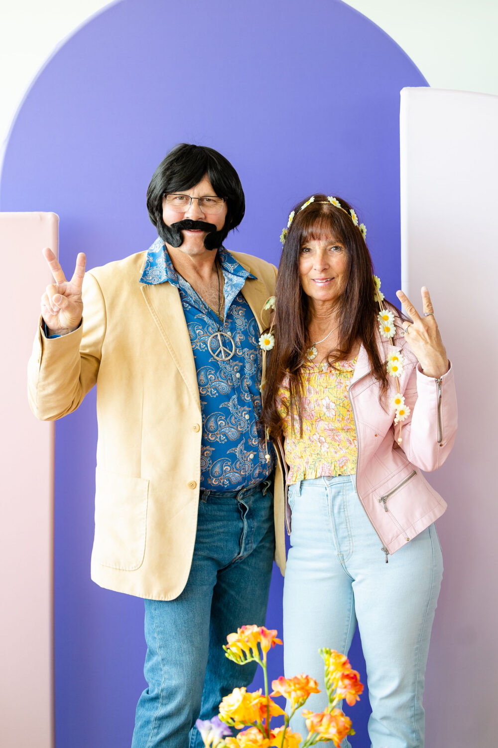 a couple (guests at a wedding) takes a photo in front of a purple backdrop. They're wearing 1970s themed clothing and holding up peace signs.