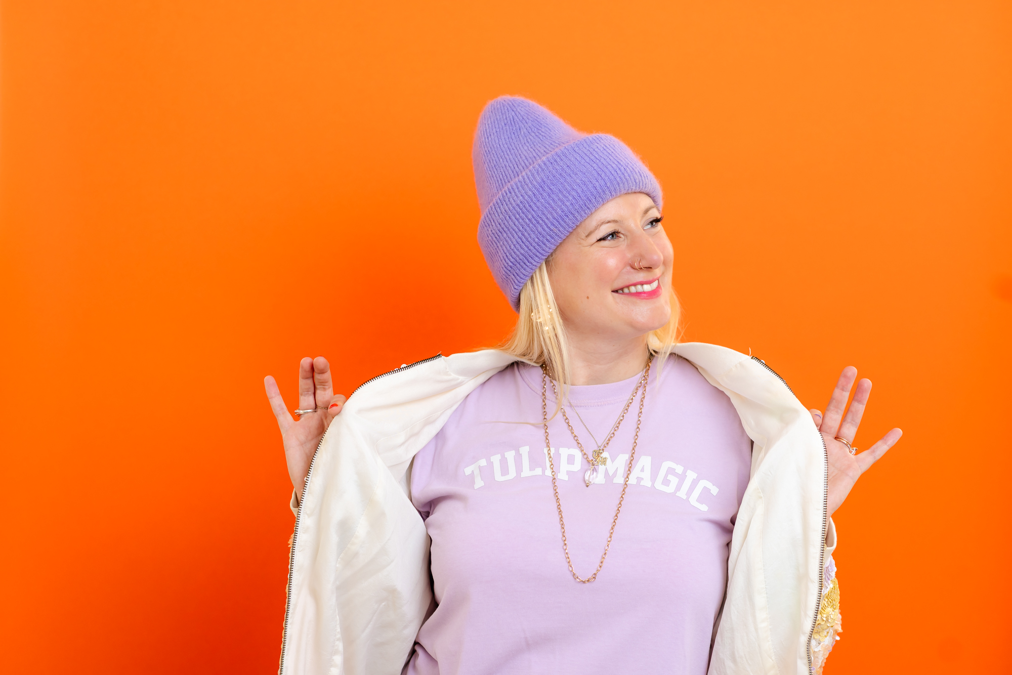 Brand photo session for a tarot card reader. She is standing in front of an orange backdrop and holding open her jacket, which shows a pink/purple shirt with the words "Tulip Magic"