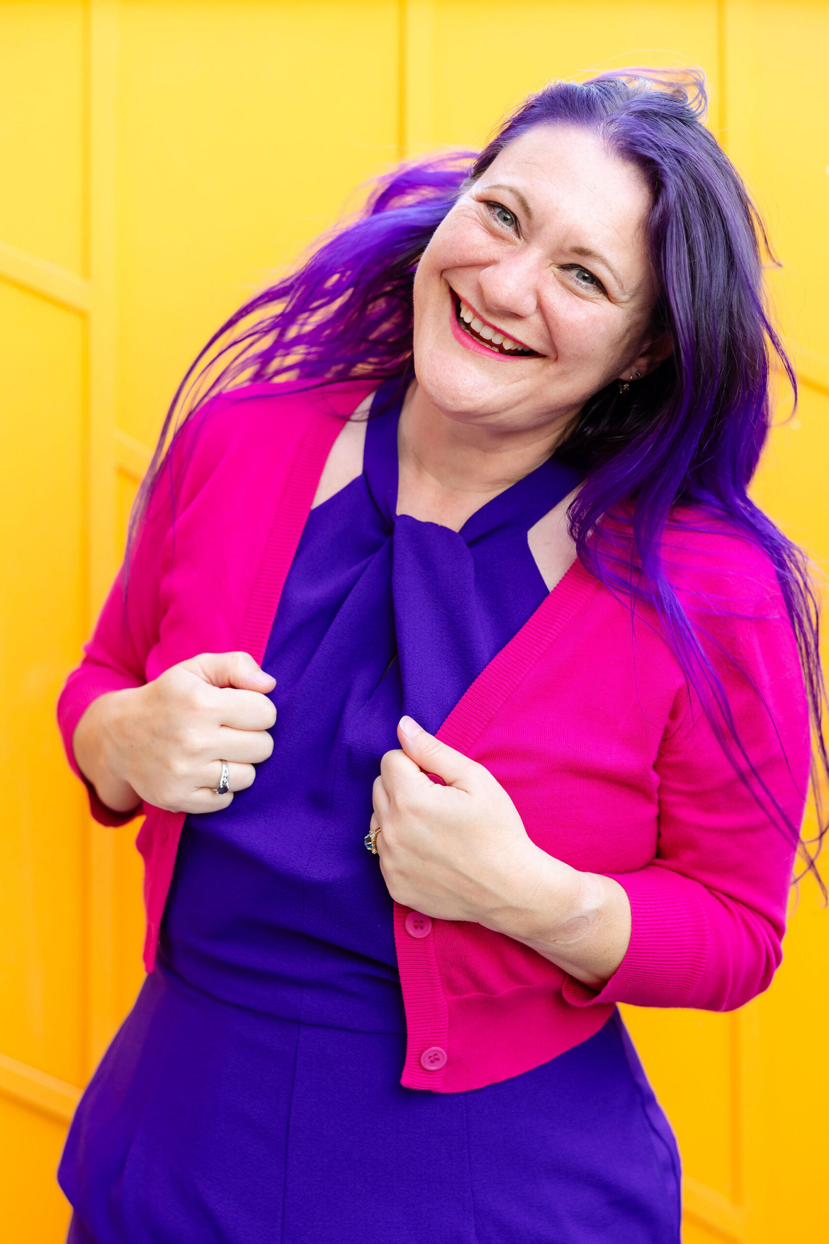 a woman with purple hair is wearing a purple jumpsuit with hot pink sweater and posing joyfully in front of an orange backdrop.