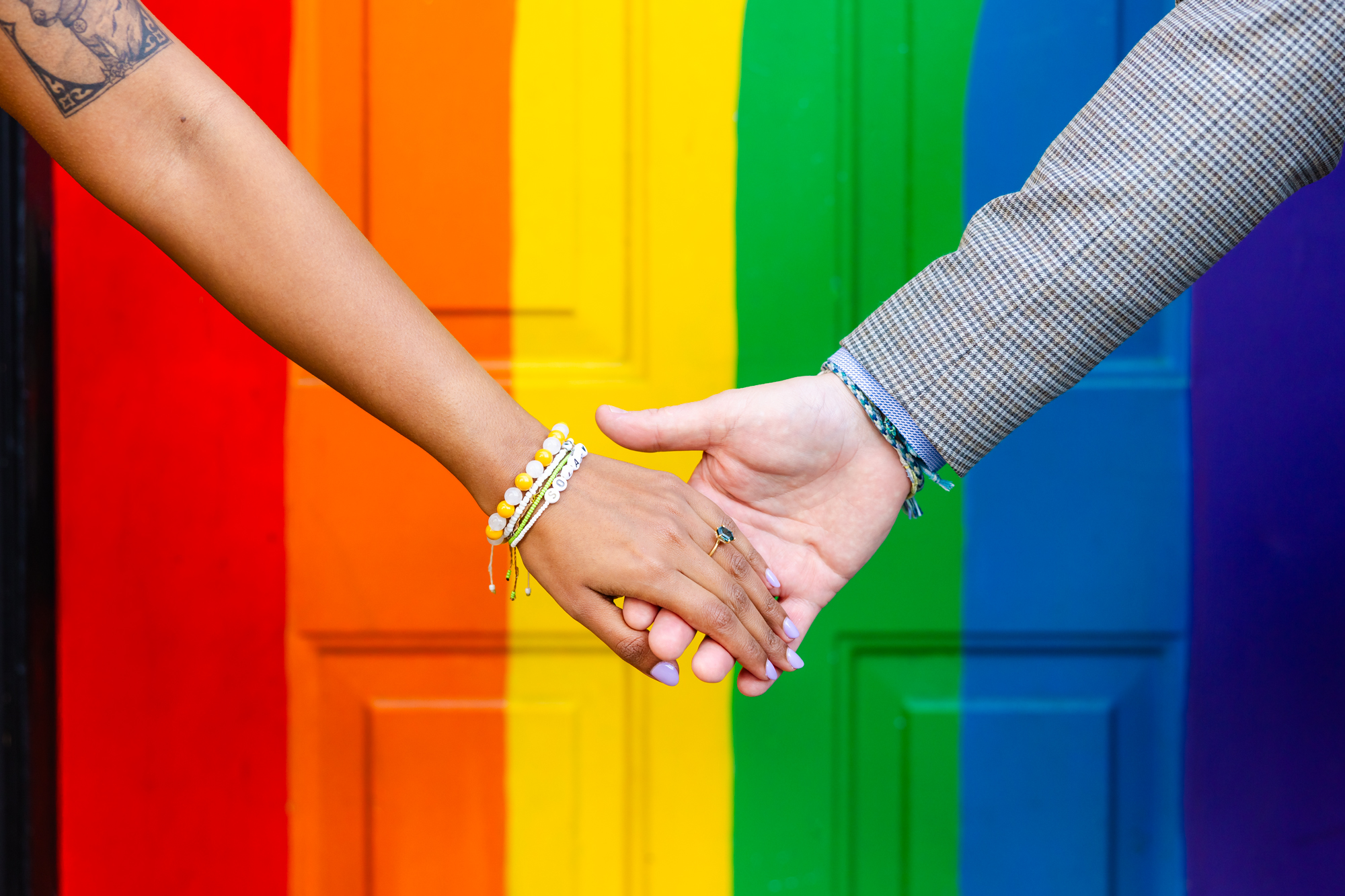Close up of a couple holding hands in front of a rainbow stripe wall.